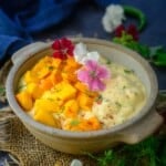 A bowl of creamy Mango Raita topped with diced mangoes, red and white edible flowers, and herbs sits on a burlap mat, with a blue cloth and more flowers in the background.
