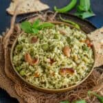 A bowl of vibrant Pudina Rice garnished with cashews and fresh mint, sits on a rustic mat with papad and green chili in the background.