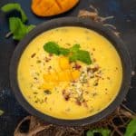 A black bowl filled with creamy Mango Basundi, is garnished with chopped mango pieces, mint leaves, and nuts on a burlap cloth. A sliced mango and fresh mint leaves rest in the background on a dark surface.