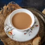 A cup of ginger chai sits on a floral-patterned saucer, placed on a piece of burlap fabric with a cookie on the side and a metal tray nearby.