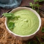 A white bowl filled with creamy Bathua Raita, garnished with fresh herbs, sits on a rustic woven mat with scattered green leaves and a blue cloth in the background.