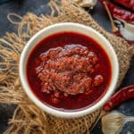 A white bowl filled with chunky garlic chutney sits on burlap, surrounded by dried red chilies and garlic cloves on a dark surface.