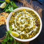 A bowl of creamy Palak Corn curry garnished with cream, served with lemon wedges and fresh herbs on a rustic mat, alongside naan bread and green chili.