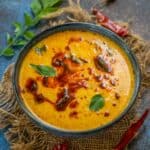 A bowl of creamy yellow Vegetable Kadhi with red chili oil, garnished with curry leaves, sits on a piece of burlap. Dried red chilies and fresh green curry leaves are scattered around on the textured surface.