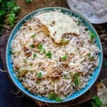 A bowl of Wadi Chawal, garnished with cilantro and a piece of papad, is served alongside creamy raita on a rustic surface with scattered herbs.