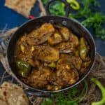 A black metal bowl filled with spicy Kalonji Baingan, surrounded by fresh herbs, whole chilies, and pieces of roti on a rustic fabric and dark blue surface.