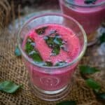A glass of pink Beetroot Chaas garnished with fresh curry leaves and a tempering of mustard seeds, placed on a rustic surface with another similar glass in the background.