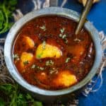 A metal bowl filled with spicy Dubki Wale Aloo curry, garnished with herbs, sits with a spoon inside. Surrounding the bowl are coriander leaves, green chili, and pieces of crispy flatbread.