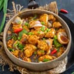 A bowl of Chili Garlic Chicken with crispy fried chicken pieces, onions, green peppers, red chilies, and savory sauce, garnished with chopped green onions.