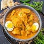 A bowl of Chicken Shahjahani curry garnished with fresh cilantro and served with halved boiled eggs. Pieces of flatbread and green chili are on the side, with some herbs scattered around on a dark surface.