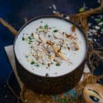 A bowl of creamy Chikoo Kheer topped with chopped nuts sits on parchment, surrounded by decorative twine and scattered nut pieces, with a halved chikoo nearby on a dark background.