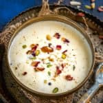 A bowl of creamy Thandai Kheer garnished with chopped nuts and dried rose petals, placed on a rustic metal tray with a spoon beside it.