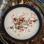 A bowl of creamy Indian Thandai Phirni garnished with sliced almonds, pistachios, rose petals, and cashews sits on a decorative metal tray with scattered nuts atop a rustic burlap mat.