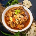 A white bowl filled with spicy Peshawari Kadhai Gosht, garnished with cilantro, sits on a rustic surface surrounded by green chilies, fresh herbs, and pieces of flatbread.