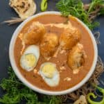 A bowl of Egg Butter Masala with halved and whole boiled eggs in a rich, orange gravy, garnished with herbs. Pieces of flatbread, fresh coriander, and green chili are placed around the bowl on a dark surface.