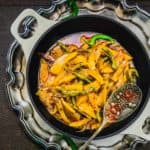 A black bowl filled with Kairi Mirch Ki Sabji, garnished with spices, sitting on an ornate silver tray with a decorative spoon beside it.