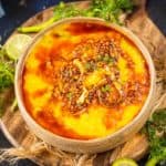 A bowl of Malwa Dal garnished with fried onions and spices, placed on a wooden board with sliced lime, green chilies, and fresh herbs around it.