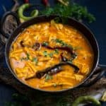 A black bowl filled with Papad Gathiya Sabji, is garnished with fresh cilantro and dried red chilies, sitting on a rustic surface with green chilies and herbs scattered around.