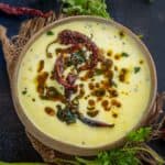 A bowl of Perugu Charu, garnished with dried red chilies, spices, and fresh cilantro, sits on a rustic cloth with green chilies and herbs scattered around on a dark surface.
