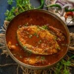 A bowl of Tomato Fish Curry in a rich red sauce, garnished with fresh cilantro. Beside it, a plate holds sliced onions, green chili, and a wedge of lime. Fresh herbs and green chilies are scattered around.