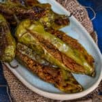 An oval plate with Besan Bhari Mirch (stuffed and roasted green chili peppers) placed on a piece of burlap, with two whole green chilies in the background on a dark blue textured surface.
