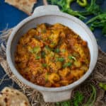 A bowl of spicy Indian Achari Turai curry garnished with fresh cilantro, surrounded by pieces of flatbread (roti) and fresh herbs, on a rustic blue surface.