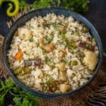 A bowl of Bengali Style Veg Pulao with visible rice, carrots, beans, cauliflower, peas, cashews, and whole spices, garnished with fresh coriander, placed on burlap with herbs and green chili peppers scattered around.