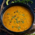 A black bowl filled with creamy orange Jaisalmeri Chana curry, garnished with chopped fresh cilantro, sits on a dark surface surrounded by green chilies, red chilies, and fresh coriander leaves.