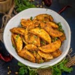 A white bowl filled with crispy, seasoned Karare Aloo wedges garnished with chopped herbs, placed on a dark table with fresh cilantro, dried red chilies, a beverage in a glass, and stacked empty glasses nearby.