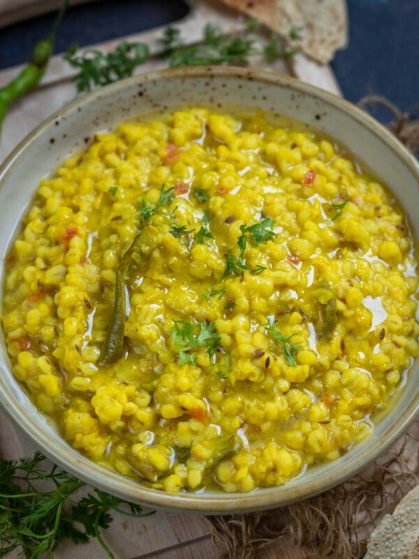 A bowl of Barley Khichdi topped with fresh herbs sits on a wooden board, surrounded by green chilies, cilantro, and crisp papad pieces, set against a rustic blue background.