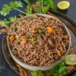 A bowl of cooked ragi vermicelli upma, featuring thin noodles, diced carrots, green beans, and peas, garnished with cilantro and surrounded by lime slices and fresh cilantro leaves on a dark surface.