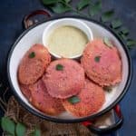 A white bowl with red handles holds five Beetroot Oats Idlis garnished with curry leaves, arranged around a small bowl of green chutney. The bowl sits on a burlap cloth, with more curry leaves scattered nearby.
