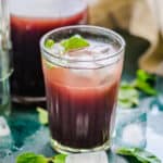 A glass of iced, dark red Kokum Juice garnished with fresh mint leaves sits on a table with scattered ice cubes and mint. A matching carafe of Kokum Juice and a beige cloth are blurred in the background.