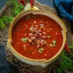 A bowl of Kashmiri Rajma curry garnished with fresh cilantro, placed on a rustic burlap mat with lemon wedges and red chili peppers nearby.