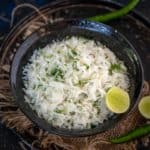 A black bowl filled with Mooli Lachha, garnished with chopped herbs and a lime wedge, placed on a rustic mat with green chilies and lime in the background.