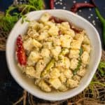 A bowl of Til Wale Aloo garnished with sesame seeds, red and green chilies, and fresh cilantro, set on a rustic surface with scattered chilies and herbs around.