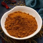 A white bowl filled with a heap of reddish-brown Achari Masala powder is placed on a dark surface, surrounded by dried red chilies, coriander seeds, a blue cloth, and jute fibers.