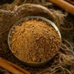 A bowl filled with Mumbai Sandwich Masala Powder, sits on a rustic tray, surrounded by coarse twine and two whole cinnamon sticks.