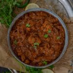 A bowl of rich, reddish-brown Achari Gobhi curry garnished with fresh cilantro leaves, surrounded by herbs and lemon slices on a rustic surface.