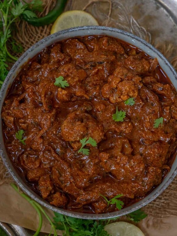 A bowl of rich, reddish-brown Achari Gobhi curry garnished with fresh cilantro leaves, surrounded by herbs and lemon slices on a rustic surface.