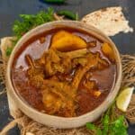 A bowl of spicy Aloo Gosht mutton curry with potatoes, garnished with herbs, served alongside pieces of flatbread, green chilies, lemon wedge, and fresh cilantro on a dark surface.