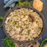 A bowl of Brown Rice Pulao garnished with fresh herbs, featuring visible peas, cauliflower, and a cinnamon stick, surrounded by papad, lime wedges, green chili, and a blue cloth on a dark surface.