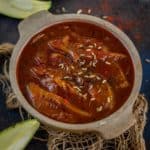 A bowl of spicy Aam ki Launji, with mango slices in a rich, red sauce, garnished with fennel and nigella seeds, sits on rustic fabric with raw mango pieces scattered around.