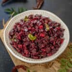 A white bowl filled with Beetroot Poriyal, garnished with curry leaves, sits on brown paper with dried red chilies and fresh curry leaves scattered around on a dark surface.