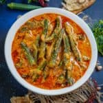 A white bowl filled with Bhindi ka Salan, an okra curry in a rich orange-red gravy, sits on a dark surface, surrounded by pieces of flatbread, dried red chilies, peanuts, garlic, fresh herbs, and okra.