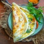 A crispy Egg Dosa served on a green plate with red chilies, cilantro, and a small bowl of podi on the side.