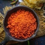A black bowl filled with red Vada Pav Chutney powder is placed on burlap, with two pav buns and green chilies in the background.