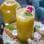 A tall glass of yellow-green Aam Panna drink with ice cubes, garnished with a sprig of mint and pink edible flowers, set on a rustic surface with scattered mint leaves and ice.