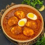 A bowl of spicy Chettinad Egg Curry with six boiled eggs in a rich red sauce, garnished with fresh coriander, placed on a dark surface with green chili, lemon wedges, and coriander leaves beside it.