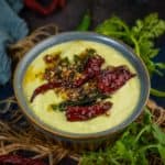 A bowl of Coconut Cilantro Chutney topped with fried red chilies, curry leaves, and spices, surrounded by fresh coriander and dried red chilies on a dark rustic surface.